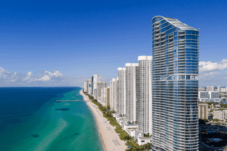 miami-condo-insurance-appraisal-reserve-study - FPAT - FPAT Aerial View of the Modern Curved Glass Ritz Carlton Residences Luxury Condominium Tower on the Beach in Sunny Isles Beach Florida with Turquoise Atlantic Ocean Waters Sandy Shoreline and a Row of Oceanfront High Rises Stretching South Under a Clear Blue Sky